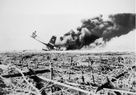 An aircraft hit by gunfire burning on the Mường Thanh airfield, viewed through a field of barbed wire.